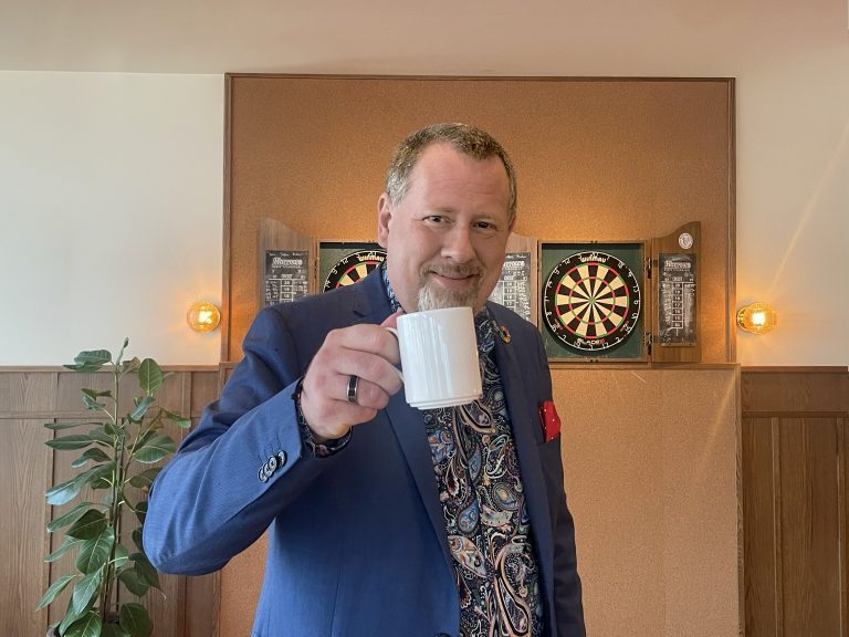 Ulf Persson skålar med en kaffekopp framför darttavlor på Hjortviken.